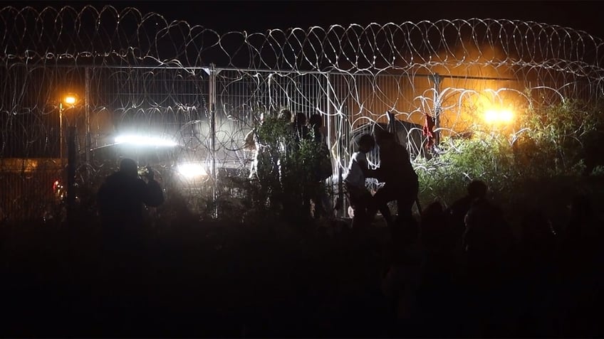 floodlights shine on border fence at night as migrants attempt crossing