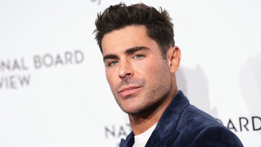 Zac Efron looks slightly back at the camera in a navy velvet suit on the carpet