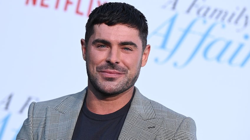 Zac Efron soft smiles on the carpet wearing a black shirt and black and white houndstooth jacket