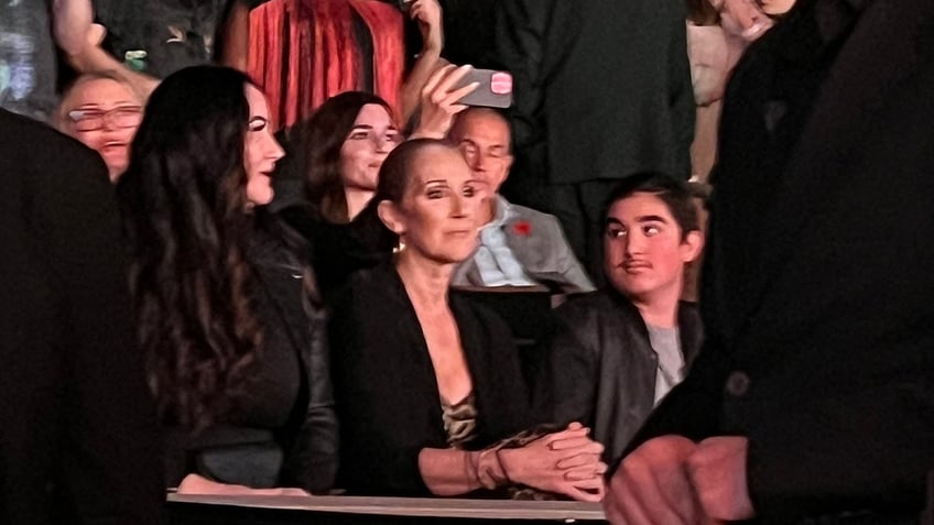 Celine Dion in a black suit seated watches Katy Perry perform in Las Vegas