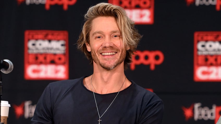 Chad Michael Murray in a black t-shirt and silver cross smiles as he sits down on stage