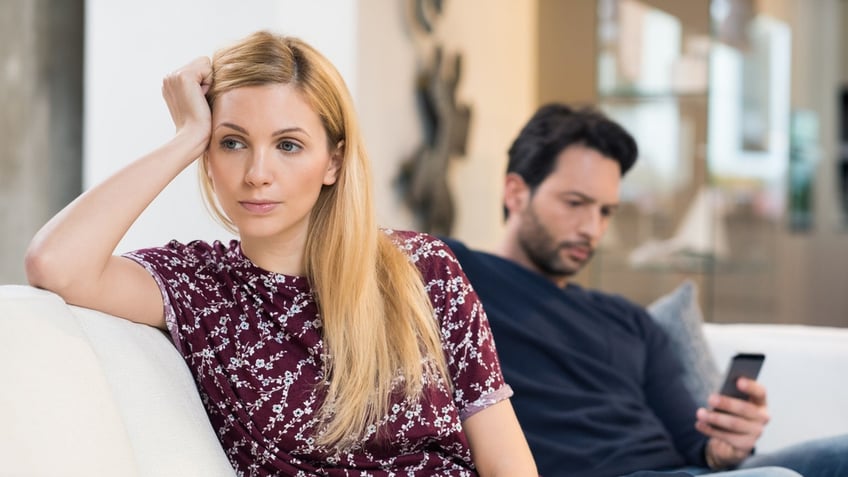 Young woman getting bored while man using phone in the background. Beautiful young woman feeling annoyed as man texting on phone. Young woman after an argument with her boyfriend in their living room.