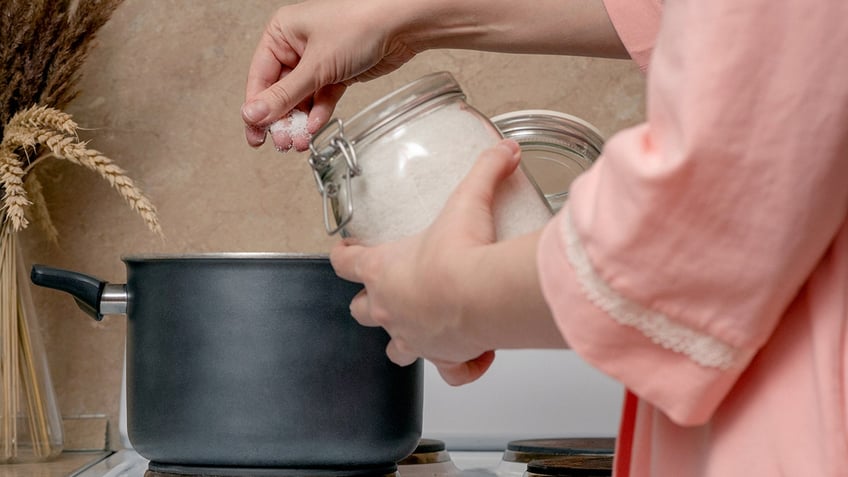 Woman is cooking food on the electric stove on home kitchen and adds salt to a saucepan.