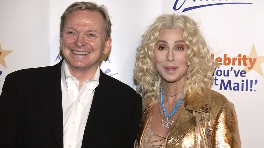 Bob Mackie in a black blazer and white shirt smiling next to Cher wearing a gold dress and a blue beaded necklace.