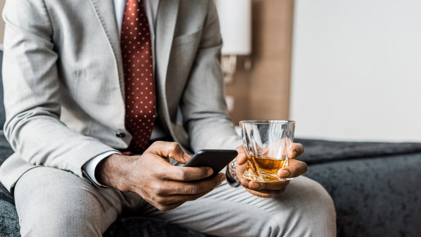 A man holds a cellphone in one hand and a glass of whiskey in another while sitting on a bed in a hotel room.