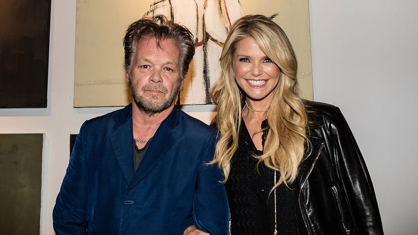 Christie Brinkley in a black leather jacket smiles next to John Mellencamp in a blue blazer