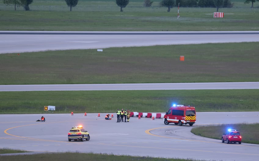 18 May 2024, Bavaria, Munich: Police and firefighters stand on a runway access road at Franz-Josef-Strauß Airport around climate activists who have stuck themselves there. Climate protection activists paralyzed Munich Airport early Saturday morning. The activists had reached the inner area of the airport grounds. According to their own statements, members of the so-called Last Generation had planned to enter the airport grounds in order to block at least one of the two runways. Photo: Karl-Josef Hildenbrand/dpa (Photo by Karl-Josef Hildenbrand/picture alliance via Getty Images)