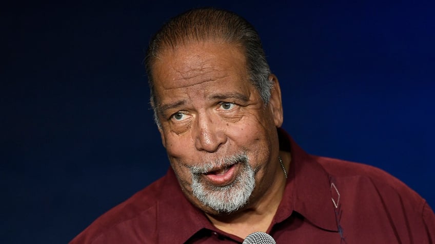 Rudy Moreno in a red shirt holding a microphone looks to his right