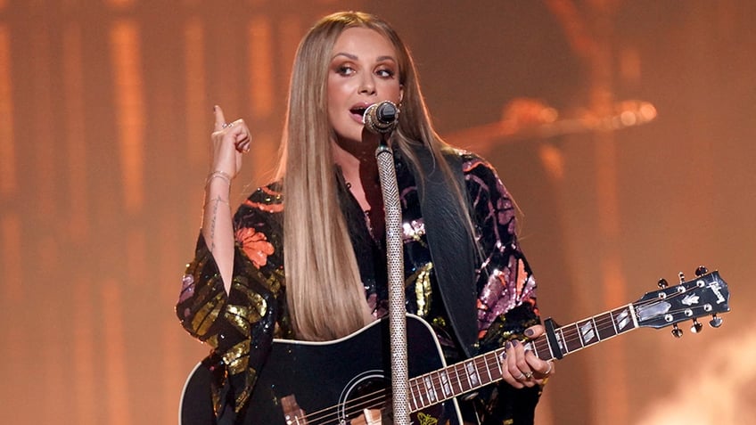 Carly Pearce in a printed sparkly top points her finger in the air and looks to her right on stage while performing at the Peoples Choice Country Awards