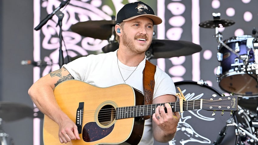 Corey Kent in a white t-shirt and black hat smiles as he looks to his left and plays the guitar on stage