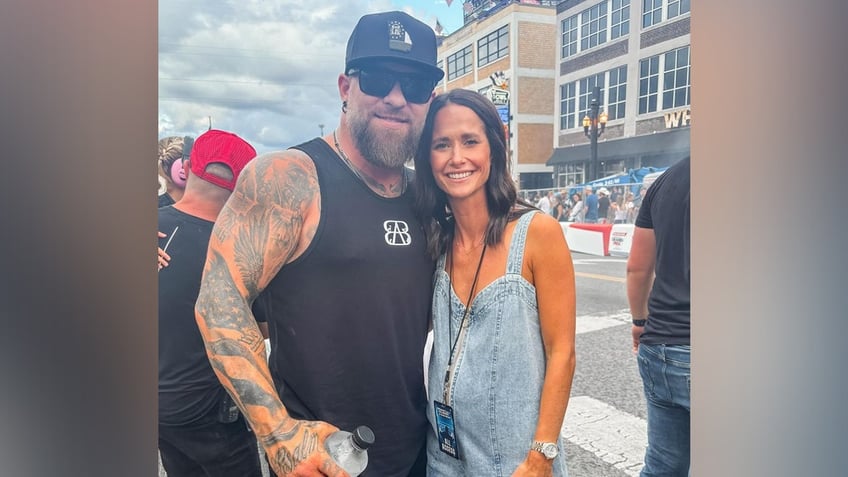 Brantley Gilbert in a black cut of shirt and hat and sunglasses smiles with his wife Amber in a denim romper