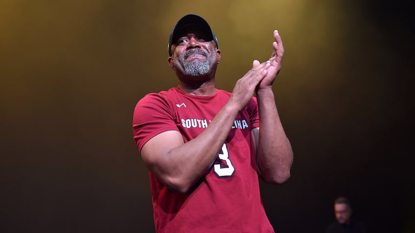 Hootie & the Blowfish singer Darius Rucker smiles on stage in a red shirt.