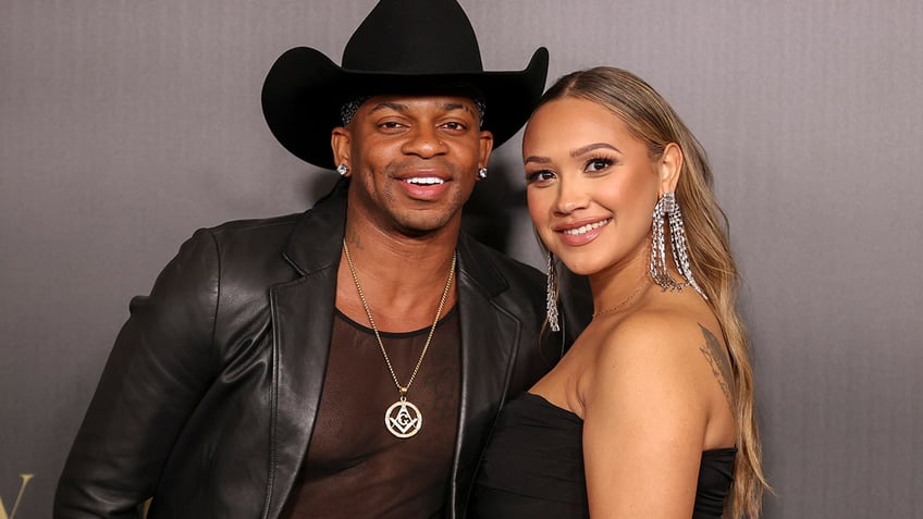 Jimmie Allen in a black leather jacket and cowboy hat poses for a photo with wife Alexis Gale