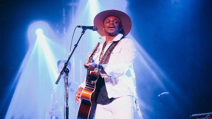 Country singer Jimmie Allen wears all white while holding a guitar.