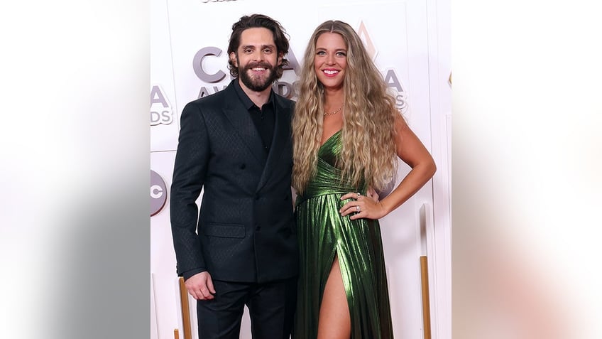 Thomas Rhett in a black suit smiles with wife Lauren with super long curly hair and a metallic green dress