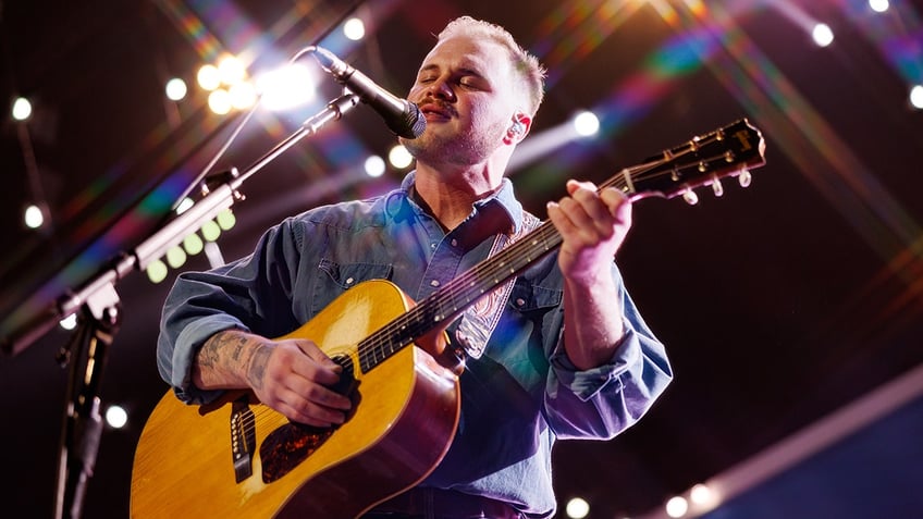 Zach Bryan in a blue denim shirt sings int o the microphone while strumming the guitar