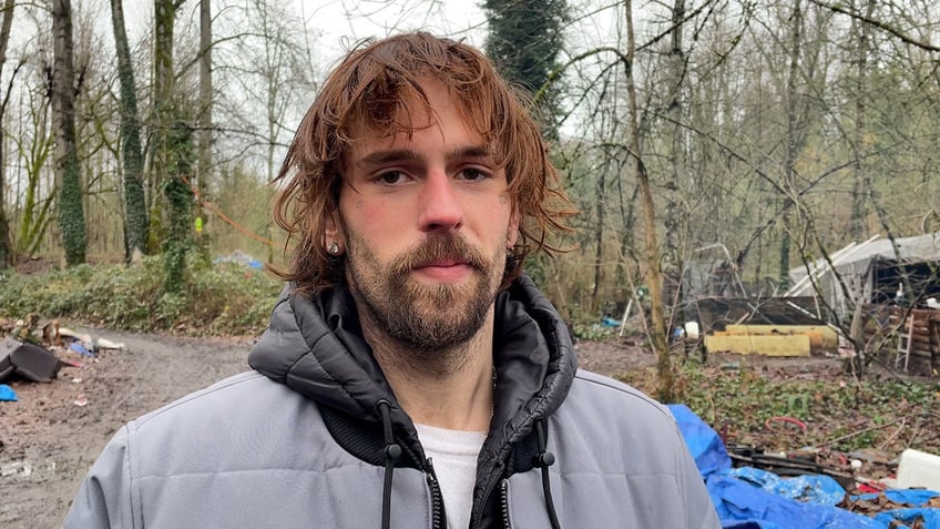 Man in gray jacket stands in homeless encampment