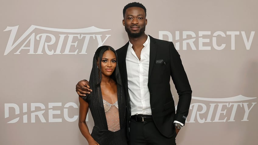 Charity Lawson in a black outfit with a sparkly bra stands next to fiancé Dotun Olubeko