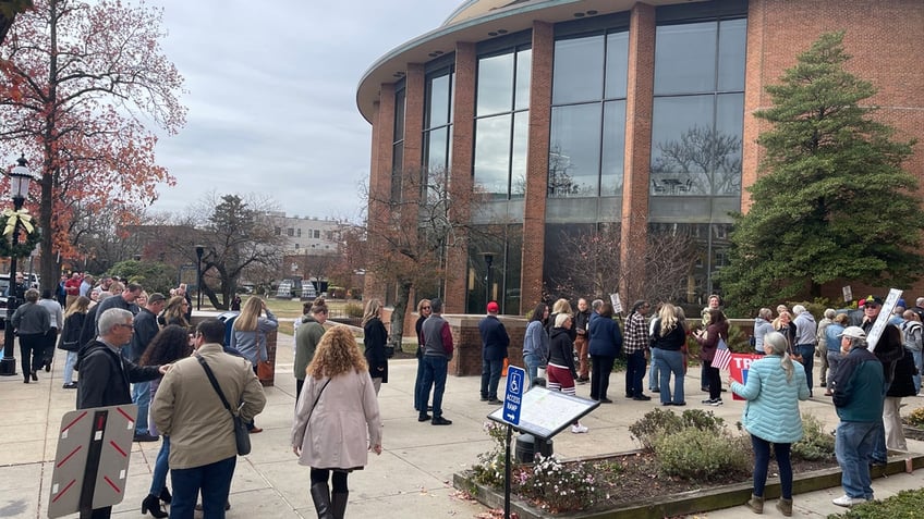 People in jackets wait in line as others join them outside of a round, brick building with large windows.