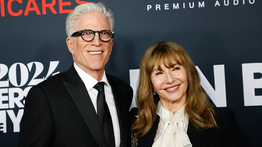 Ted Danson smiles on the carpet wearing a black suit and tie with wife Mary Steenburgen in a white blouse and black jacket