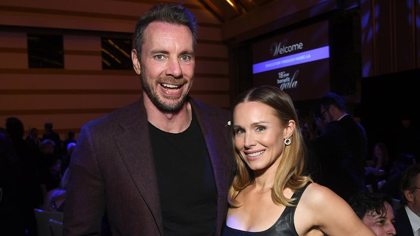 Dax Shepard smiles/laughs for a photo wearing a black shirt and purple jacket with wife Kristen Bell in a black dress