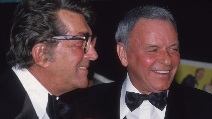 Dean Martin smiling next to Frank Sinatra as they both wear tuxes.