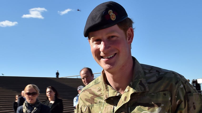 Prince Harry smiles in a black cap and camo military gear