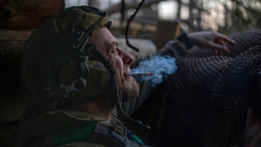 A Ukrainian serviceman from the Azov brigade, known by the call sign Chaos, smokes a cigarette in a dugout while he waits for a command to fire