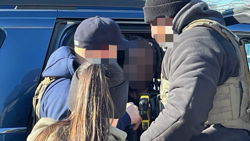 Three ICE officers stand around a man in a police car.