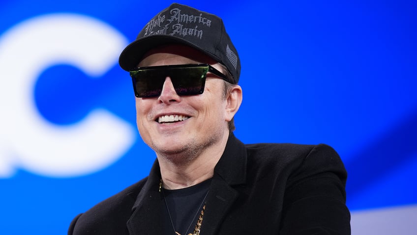 Billionaire Elon Musk pictured in a hat and sunglasses, smiling. 