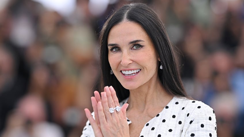 Demi Moore smiles and clasps her hands together in a white dress with black polka dots at Cannes