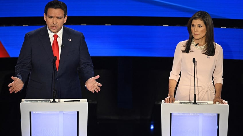 Ron DeSantis wearing navy suit, bright red tie, with arms out and hands open, talking (left), Nikki Haley wearing light pink dress, pearl necklace serious expression (right)