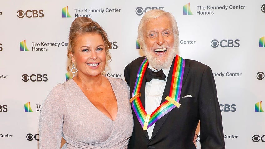Dick Van Dyke in a tuxedo and rainbow medal smiles with his wife Arlene in a light purple dress