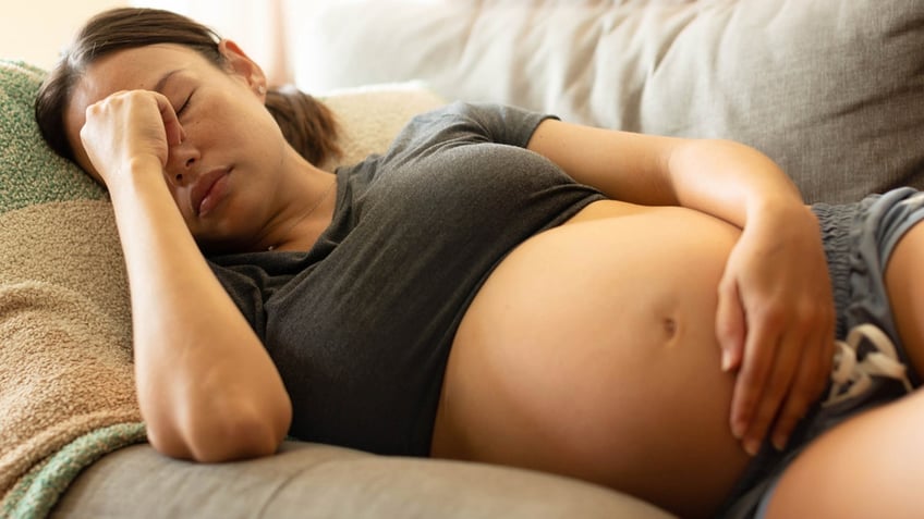 A uncomfortable pregnant mother trying to rest at home, lying on her couch holding her head.