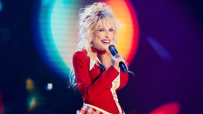 Dolly Parton in a red shirt smiles and looks at the camera holding a microphone