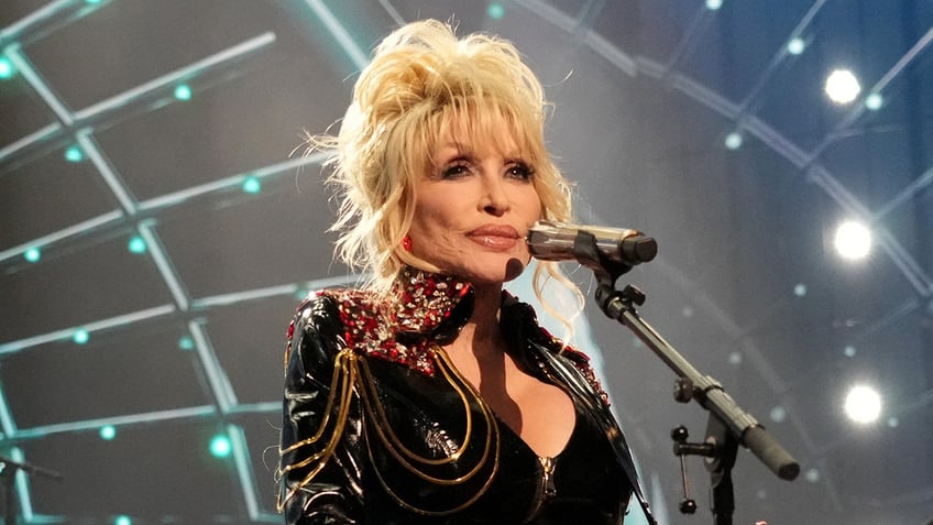 Dolly Parton in a black jacket sings on stage and looks out