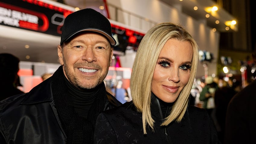 Donnie Wahlberg in a black turtleneck, scarf, jacket and hat smiles for the camera next to wife Jenny McCarthy, also wearing black