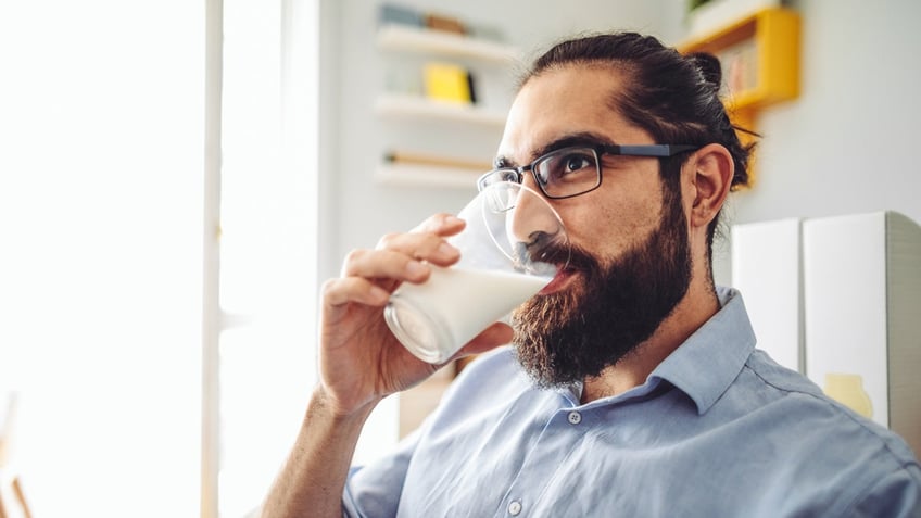 A bearded man wearing glasses drinks a glass of milk while looking in front of him.