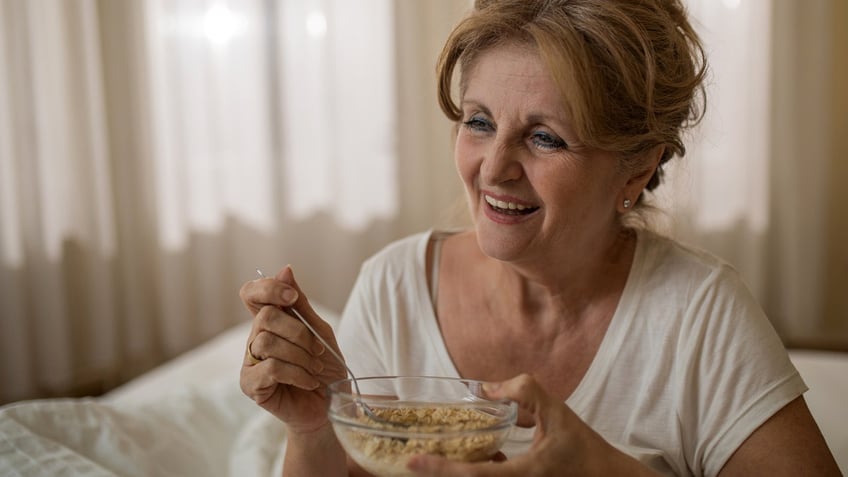 A mature woman eats oatmeal in bed while smiling.