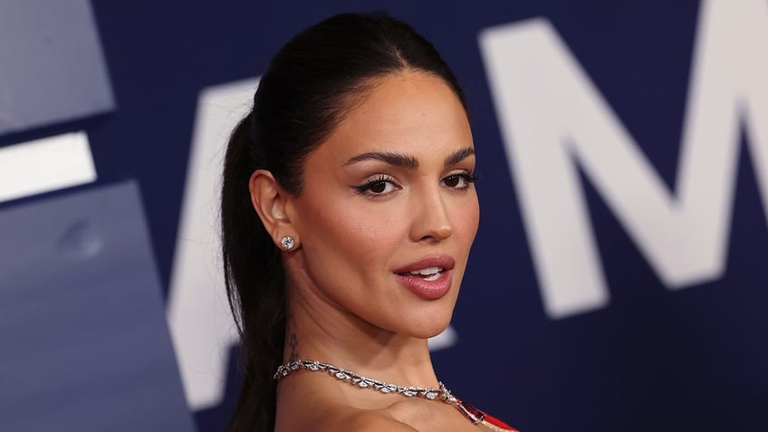 Eiza González with her hair in a ponytail wearing a red dress and diamond stud earrings poses on the carpet