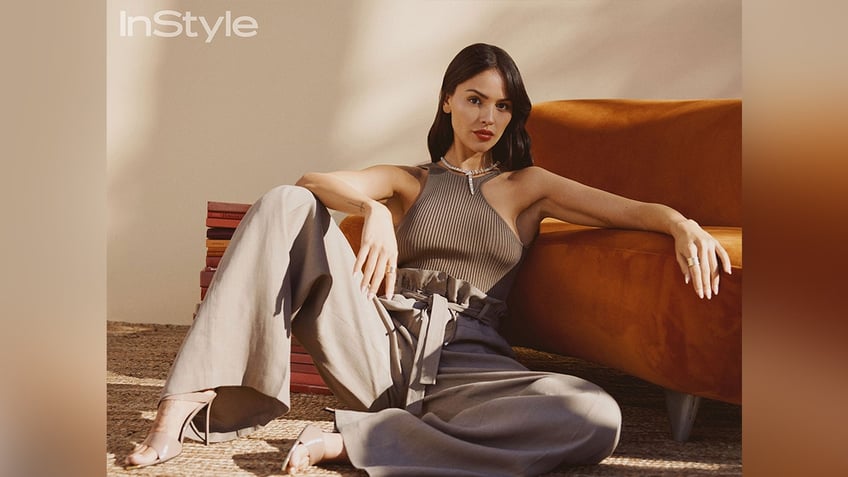 Eiza González leans against a burnt orange couch in a brown ribbed tank top and light colored slacks