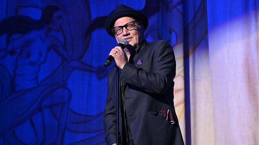 Rob Schneider in a top hat and navy suit performs on stage