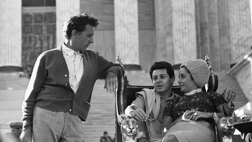 Richard Burton looking at Elizabeth Taylor and Eddie Fisher as they sit together.