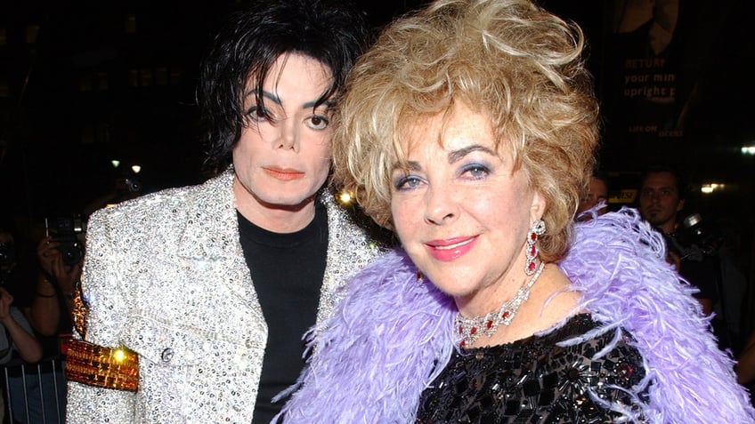 A close-up of Michael Jackson wearing a silver blazer and black shirt next to Elizabeth Taylor wearing a sparkly black dress and a purple boa.