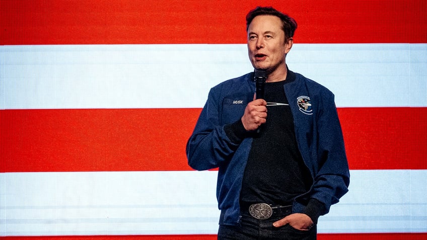 Elon Musk speaking at event with an American flag in background