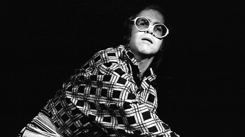 Elton John looking confused while wearing a multiprinted shirt