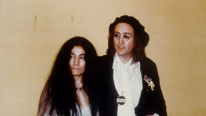 Yoko Ono pursing her mouth as she stands next to John Lennon who looks on.