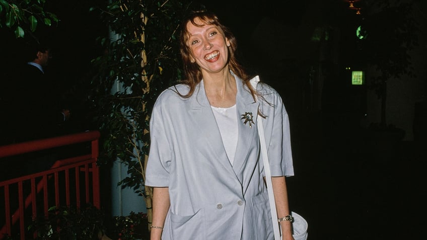 Shelly Duval smiles in a light blue/grey quarter-length blazer