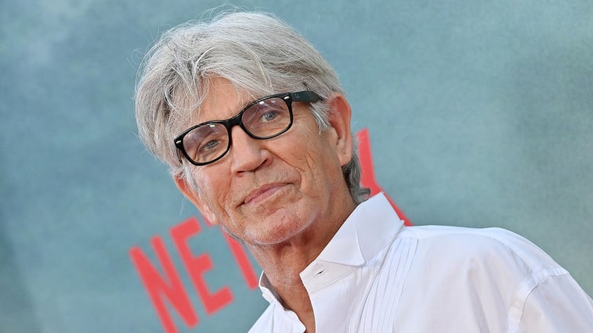 Eric Roberts wears black frame glasses and white shirt on the carpet