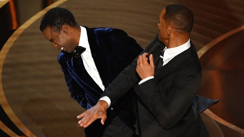 Chris Rock in a velvet tuxedo winces from impact after Will Smith in a black tuxedo slaps him across the face